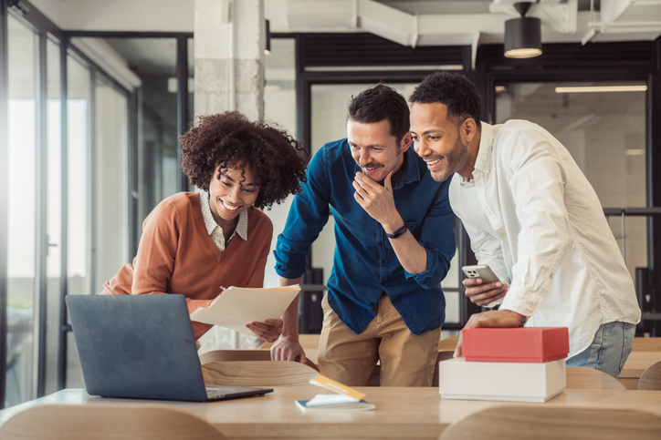 Three colleagues smiling and collaborating around a laptop in a modern office, reviewing documents and working together.