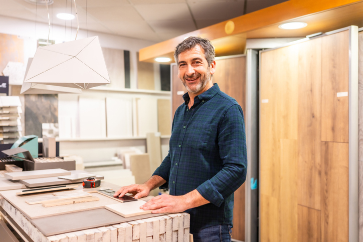 A smiling man stands behind a retail counter in a flooring showroom, surrounded by wood flooring samples, tiles, and design boards. The store has modern lighting and a variety of floor products displayed vertically behind him.
