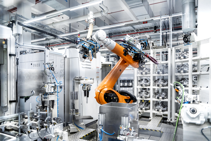 Robotic arm operating in a high-tech automated manufacturing facility surrounded by machinery and component trays.