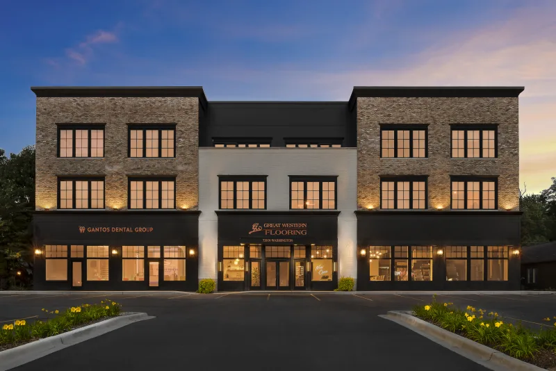 Exterior view of the Great Western Flooring storefront at dusk, featuring warm lighting, a three-story brick-and-stucco building, and adjacent signage for Gantos Dental Group.          Ask ChatGPT